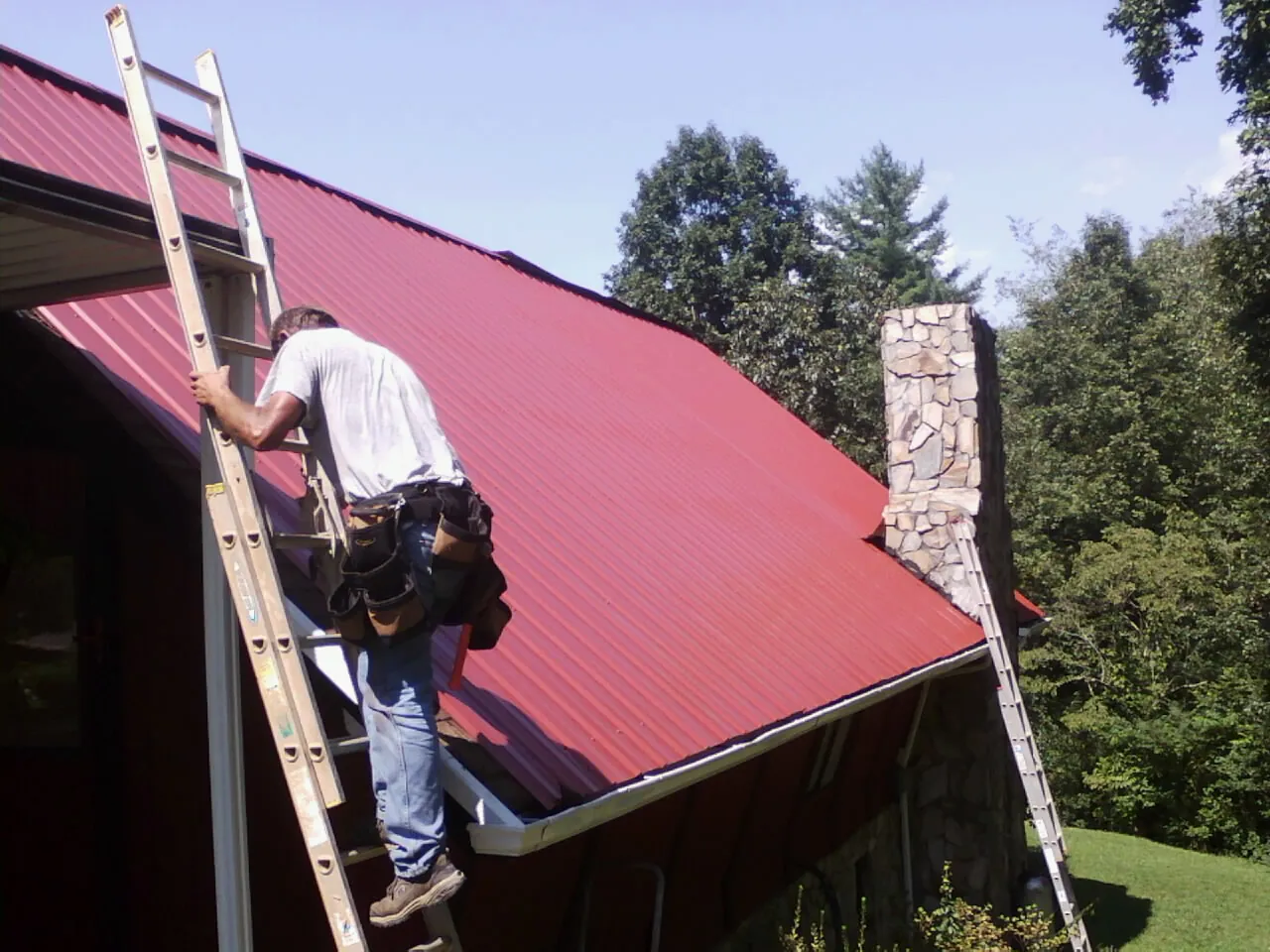 Roofer on a ladder beside a red metal roof and stone chimney — Soffit & Fascia Repair by Roofing Brothers in Huntsville